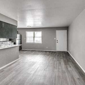 Empty living room with gray plank flooring opens to a kitchen with white counters and stainless appliances