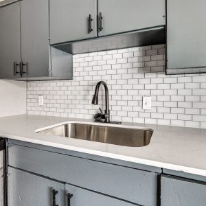 Kitchen with gray cabinets, white subway tile backsplash, quartz counter, and black faucet over stainless sink